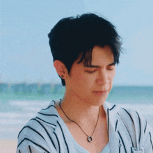 a young man wearing a striped shirt and a necklace is standing on a beach with his eyes closed