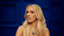 a blonde woman wearing a heart shaped necklace looks at the camera