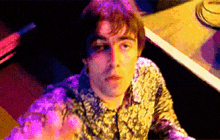 a man in a floral shirt is sitting at a table with purple lights behind him