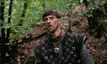 a man with a beard and chain mail is standing in the woods .