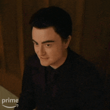 a man in a black suit and tie is sitting in front of a wooden wall with a prime arrow in the corner