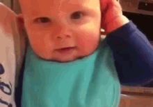 a baby is wearing a blue bib and covering his ears with his hands .