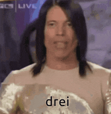 a man with long hair is wearing a white shirt and holding a trophy with the word drei written on it .