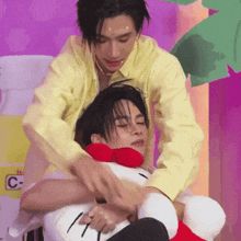 a man in a yellow shirt is hugging another man in a yellow shirt while holding a stuffed hello kitty .