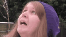 a woman wearing a purple beanie is looking up at the sky with her mouth open .