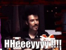 a man with a mustache is sitting at a table with a sign that says " hheeeyyy !!! "