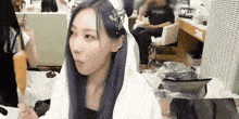 a woman with long blue hair is sitting in a room with a white blanket on her head .