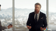 a man in a suit and tie stands in front of a window with a nbc logo on the bottom