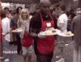 a man in a red apron is carrying plates of food in front of a crowd of people .