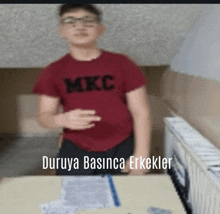 a boy wearing a red mkc shirt stands in front of a desk