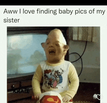 a baby with a huge head is sitting in front of a television and holding a superman hat .