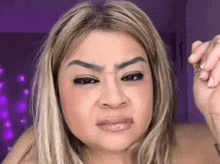 a woman with blonde hair and a purple background is making a funny face with her hand .