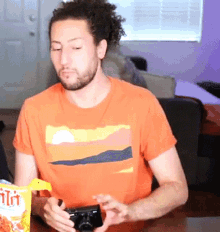a man in an orange shirt is holding a camera and a bag of fritos
