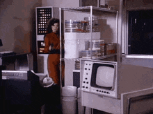 a woman is standing in a room with a television and a shelf filled with containers .