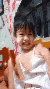 a little girl in a white tank top is smiling and looking at the camera