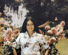 a woman in a floral shirt is standing in a garden surrounded by flowers .