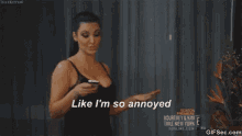 a woman in a black tank top is holding a cell phone and says `` like i 'm so annoyed '' .