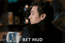 a man in a black jacket is sitting at a table with the words bet mud written on his face .