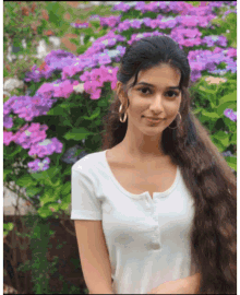 a woman in a white shirt stands in front of a bush with purple flowers
