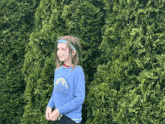 a young girl wearing a blue shirt with a rainbow on it is standing in front of a green bush