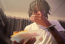 a man with dreadlocks is holding a plate of food and covering his mouth with his hand