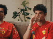 two men wearing red ferrari shirts sit on a couch drinking from glasses