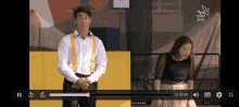 a man in a white shirt and yellow suspenders is standing next to a woman in a black dress on a video screen