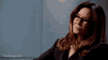 a woman wearing glasses and a black jacket is sitting in front of a wall .