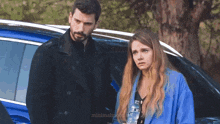 a man and a woman are standing in front of a blue car and the woman is holding a bottle of water ..