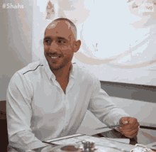 a man in a white shirt is sitting at a table with a fork in his hand and smiling .