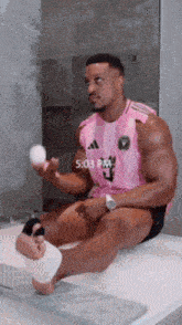 a man in a pink adidas shirt is sitting on a bathtub holding an egg