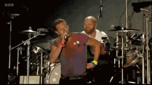 a man is singing into a microphone on a stage while a drummer plays drums .