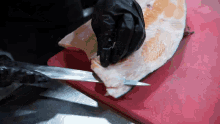 a person wearing black gloves is cutting a fish on a red cutting board