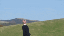 a man with pink hair stands in a grassy field with mountains in the background