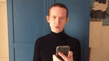 a woman in a black turtleneck holds a cell phone in front of a blue door