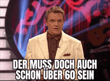 a man in a white suit and bow tie is standing in front of a sign that says der muss doch auch schon über 60 sein