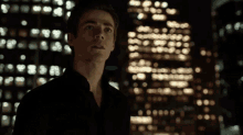 a man in a black shirt stands in front of a blurry city skyline and the word cool is on the screen