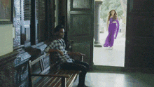 a man sits on a bench while a woman in a purple dress walks past him