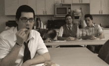 a man wearing glasses sits at a table eating a snack