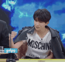 a young man wearing a moschino t-shirt is sitting at a table .