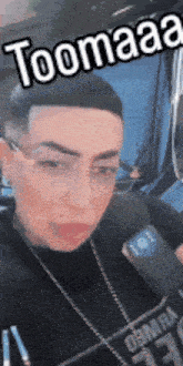 a man wearing glasses and a black shirt is talking into a microphone while sitting in a car .