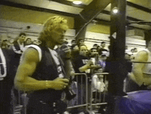 a man in a black vest with the letter e on it stands in front of a crowd holding a camera .