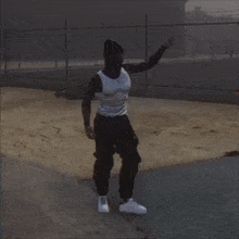 a man wearing a white tank top and black pants is standing on a dirt road .