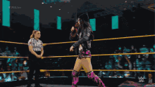 a woman in a black jacket and pink shorts stands in a wrestling ring next to a referee