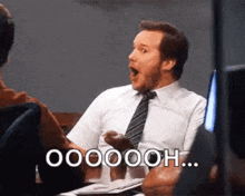 a man in a white shirt and tie is sitting at a table with his mouth open and says oooooooh .