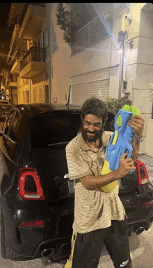 a man with a beard is holding a large water gun in front of a black car