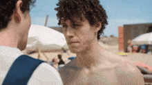 a shirtless man with curly hair looks at another shirtless man