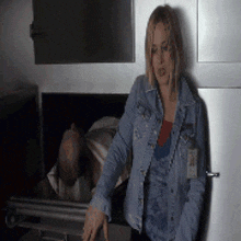 a woman in a denim jacket is standing next to a dead man in a coffin