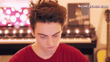 a young man in a red shirt is sitting in front of a piano making a funny face .