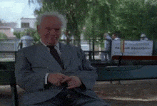 a man in a suit and tie sits on a bench in front of a sign that says parque saavedra